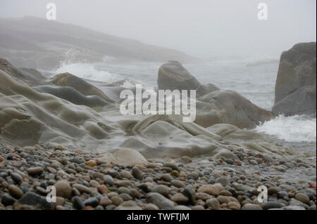 A smoothly worn rocky coastline with crashing waves and small pebbles on a foggy day. Stock Photo
