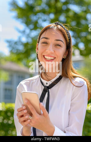 Happy vivacious young woman with a beaming friendly smile holding her mobile phone outdoors in spring sunshine Stock Photo