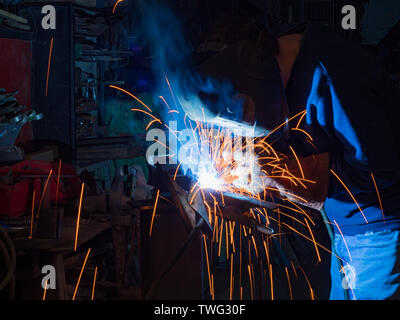 Metal working mechanic fabricator Technician MIG welding in a dark workshop with sparks flying shrouded in fumes Stock Photo