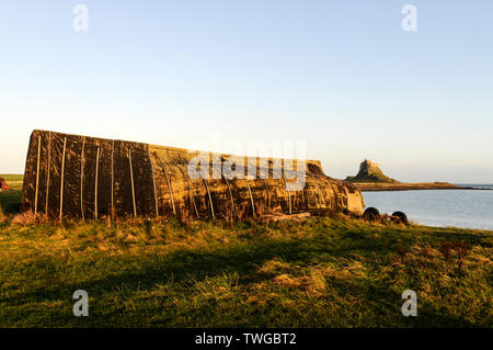 An old fishing boat turned upside down and used as a boat storage hut on the beach at Lindisfarne Holy Island with Lindisfarne Castle in the Stock Photo