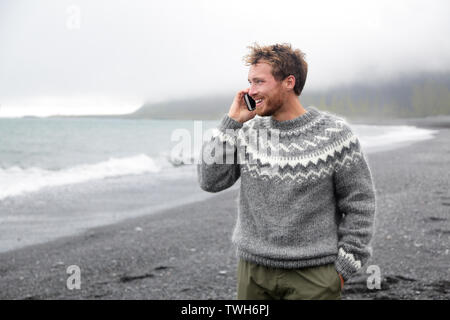 Smartphone man talking on smart phone walking on black sand beach on Iceland wearing Icelandic sweater. Stock Photo