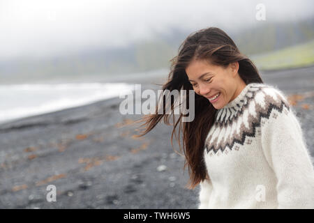 Cute woman walking on black sand beach on Iceland wearing Icelandic sweater. Pretty beautiful adorable multiracial Asian / Caucasian female model looking shy down by the ocean sea smiling happy.