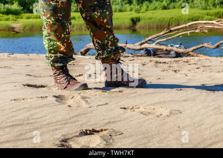 men's feet in boots and camouflage pants on the sand by the river Stock Photo