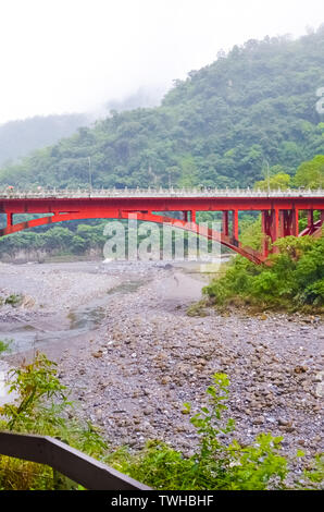 Vertical picture of red bridge in Taiwanese Taroko National Park. Taroko Gorge is a popular tourist attraction. River surrounded by green hilly landscape, forests and rocks. Misty fog, rainy weather. Stock Photo