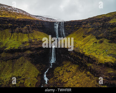 Aerial footage of Fossa waterfall from above during early spring with snow-covered mountain peaks and lush green tones (Faroe Islands, Denmark)