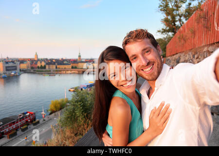 Couple taking selfie self portrait in Stockholm. Candid fresh Scandinavian man and Asian woman looking at old town cityscape sunset view from Monteliusvagen overlooking Gamla Stan, the old city. Stock Photo
