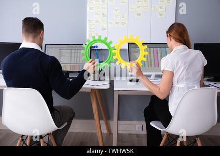 Rear View Of Two Businesspeople Joining Green And Yellow Gears While Working On Computer Stock Photo