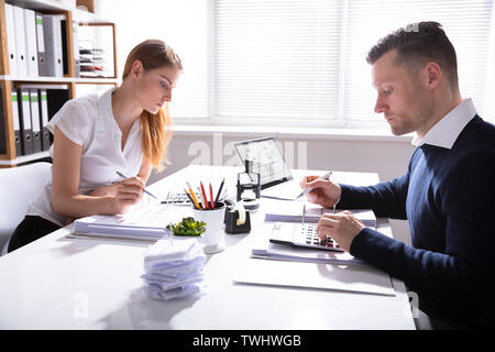 Side View Of Businessman And Businesswoman Calculating Invoice With Calculator Over White Desk In Office Stock Photo
