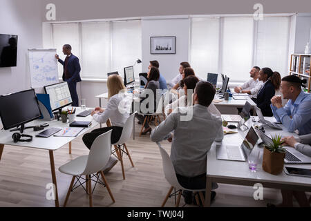 African Businessman Giving Presentation To His Colleagues Sitting On Table With Computers In Office Stock Photo