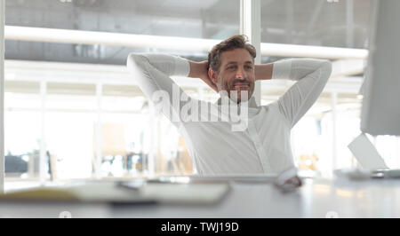 Business male executive with hands behind hand sitting at desk in a modern office Stock Photo