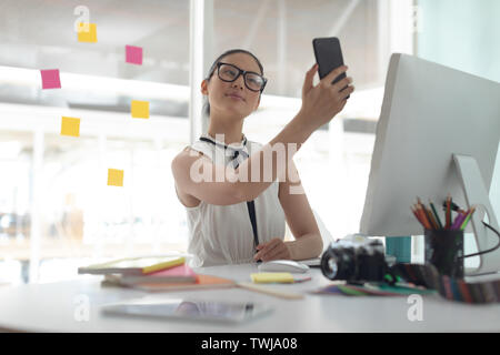 Female graphic designer taking selfie with mobile phone at desk in a modern office Stock Photo