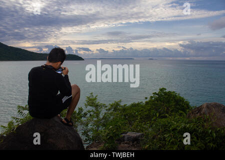 A photographer is enjoying the scenery on an island and the sea from a hill. He captured the moment using his DSLR camera Stock Photo