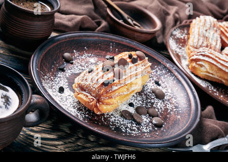 delicious Homemade Eclairs sprinkled with powdered sugar and chocolate drops on an earthenware plate on a dark rustic old table with cup of coffee, ho Stock Photo