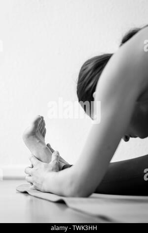 Close-up side view of a yogini. A young adult woman doing forward bending Head-to-Knee Forward Bend, or Janu Sirsasana yoga pose, black and white Stock Photo