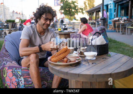 Ukraine, Kyiv, 20.06.2019. day of argentinian flag.Middle eastern man eating breakfast at outdoor restaurant. Arab man listening to music in headphones Stock Photo