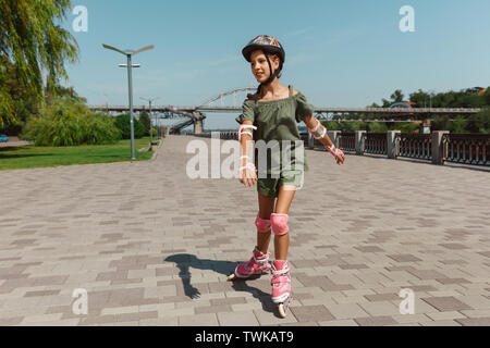 Teenage girl in a helmet learns to ride on roller skates holding a balance or rollerblading and spin at the city's street in sunny summer day. Healthy lifestyle, childhood, hobby, leisure activity. Stock Photo