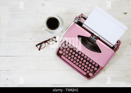 Old pink typewriter from the 1950s with blank paper, coffee and glasses on white painted rustic wood, copy space, high angle view from above, selected Stock Photo