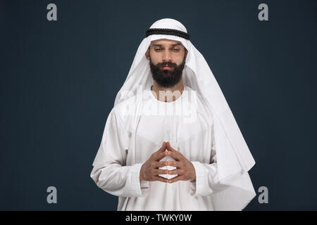 Half-length portrait of arabian saudi businessman on dark blue studio background. Young male model praying and looks thoughtful. Concept of business, finance, facial expression, human emotions. Stock Photo