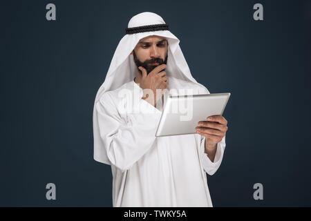 Half-length portrait of arabian saudi businessman on dark blue studio background. Young male model using tablet or gadget. Concept of business, finance, facial expression, human emotions, technologies. Stock Photo