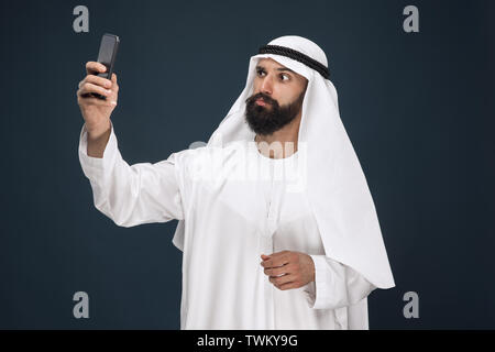 Half-length portrait of arabian saudi man on dark blue studio background. Young male model using smartphone, making selfie. Concept of business, finance, facial expression, human emotions, technologies. Stock Photo