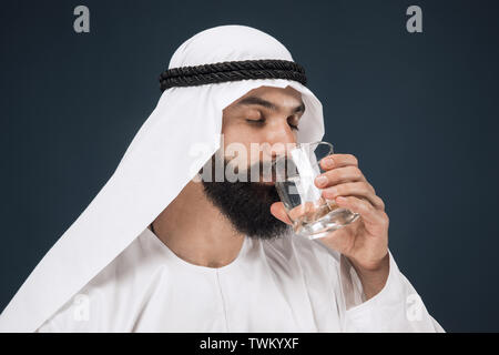 Half-length portrait of arabian saudi businessman on dark blue studio background. Young male model standing and drinking water. Concept of business, finance, facial expression, human emotions. Stock Photo