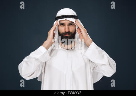 Half-length portrait of arabian saudi businessman on dark blue studio background. Young male model standing and suffering from headache. Concept of business, finance, facial expression, human emotions. Stock Photo