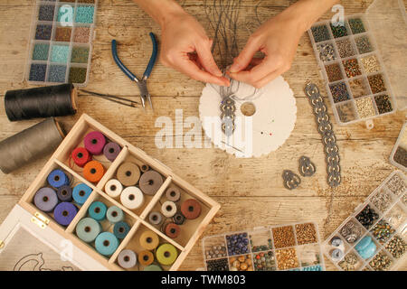 Top view of female hands making a macramé bracelet with kumihimo on a wooden table with tools, spools of thread, natural stones and colored beads Stock Photo