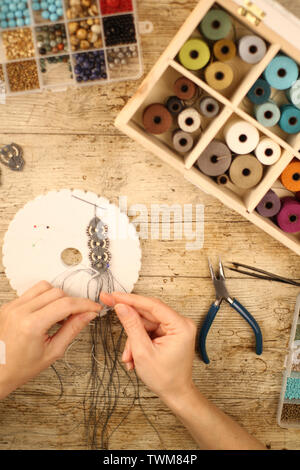 Top view of female hands making a macramé bracelet with kumihimo on a wooden table with tools, spools of thread, natural stones and colored beads Stock Photo