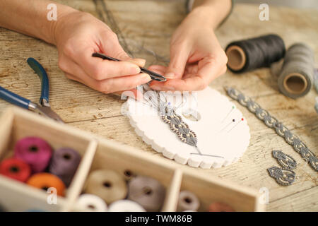 Close up of female hands making a macramé bracelet with kumihimo on a wooden table with tools, spools of thread, natural stones and colored beads Stock Photo