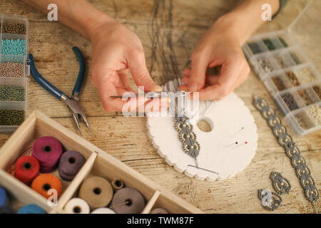 Close up of female hands making a macramé bracelet with kumihimo on a wooden table with tools, spools of thread, natural stones and colored beads Stock Photo
