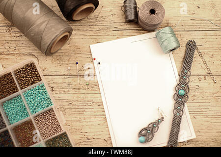 Top view of a wooden table with tools, spools of thread, natural stones and colored beads to make macramé jewelry, with blank sheet for copy space Stock Photo