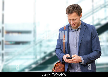Young urban professional man using smart phone in office building indoors. Businessman holding mobile smartphone using app texting sms message wearing suit jacket and bag. Stock Photo