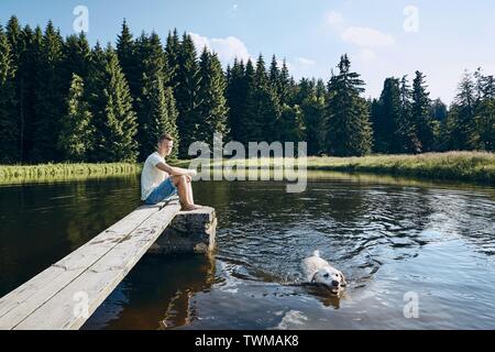 Idyllic summertime with dog. Young man looking at swimming labrador in lake against forest. Stock Photo