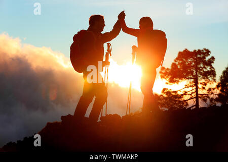 Hiking people reaching summit top giving high five at mountain top at sunset. Happy hiker couple silhouette. Success, achievement and accomplishment people Stock Photo