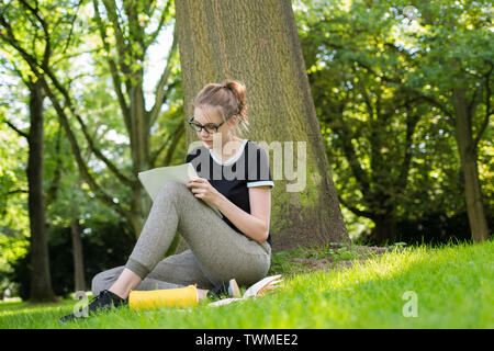 A young woman is sitting under a tree and is writing in a folder Stock Photo