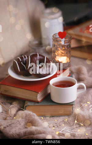 Still life details in home interior of living room. Cup of tea and chocolate donuts on stack of book. Tea leisure time in autumn and winter. Cozy rela Stock Photo