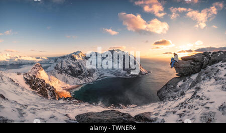 Panorama of Mountaineer sitting on rock on peak mountain of arctic coastline at sunset. Ryten Mountain, Lofoten islands, Norway Stock Photo