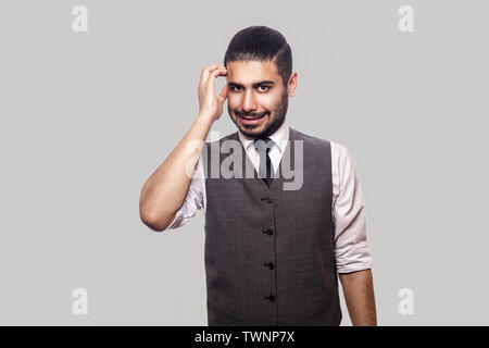 Portrait of thoughtful handsome bearded brunette man in white shirt, waistcoat standing scratching his head, looking at camera and thinking what to do Stock Photo