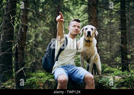 Hiker with dog in forest. Man and his labrador retriever resting on fallen tree. Stock Photo