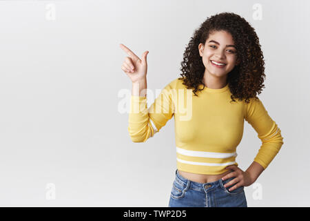 Carefree confident friendly smiling upbeat lively girl curly hairstyle pointing upper left corner hold hand waist self-assured relaxed grinning deligh Stock Photo