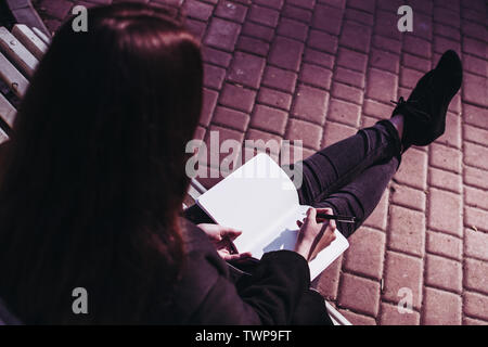 Brunette girl working on her homework assignment essay or research paper and writing down her thoughts in a notebook. Paved sidewalk on a background