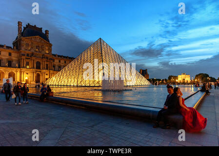 A couple taking engagement photos by the illuminated glass pyramid at the Louvre Museum in Paris, France at dusk Stock Photo