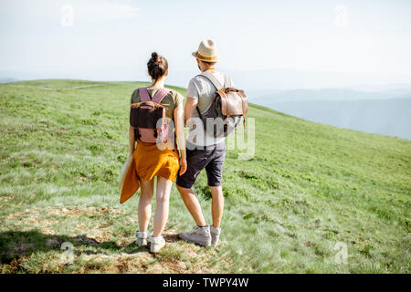 Young couple standing together with backpacks, enjoying beautiful landscape view on the green mountains, rear view Stock Photo