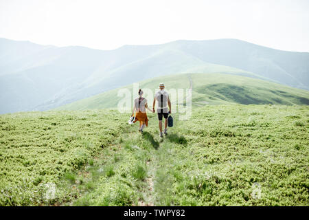 Couple walking with backpacks on the green meadow, traveling in the mountains during the summer time, wide landscape view Stock Photo