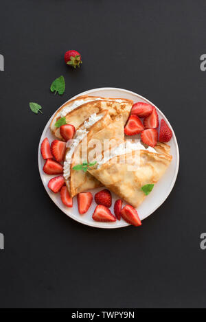 Crepes with ricotta cheese and fresh strawberries on black background, top view, copy space. Delicious crepes, thin pancakes. Stock Photo