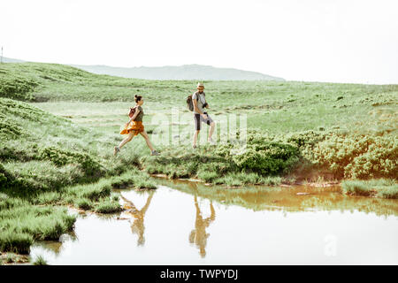 Man and woman hiking near the lake in the mountains, landscape view on the green meadow with lake during the sunny day Stock Photo