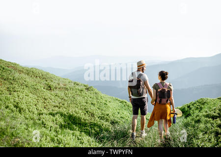 Beautiful couple walking with backpacks on the green meadow, while traveling high in the mountains during the summer time. Rear view Stock Photo