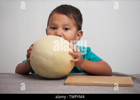 An impatient toddler trying hard to take a bite out of a honeymelon he is holding in his grasp before being served. Stock Photo