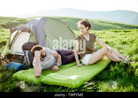 Young and cheerful couple lying on the mattress at the campsite, enjoying summer time while traveling in the mountains Stock Photo
