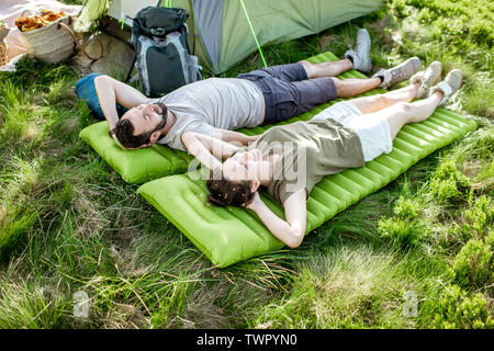 Young and cheerful couple lying on the mattress at the campsite, enjoying summer time while traveling in the mountains Stock Photo